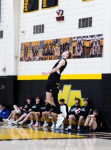 Athlete performing a volleyball serve in gym, teammates watching from the sidelines. Energetic team sport moment captured.