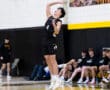 A volleyball player jumps to serve during a match, with teammates and a scoreboard visible in the background.