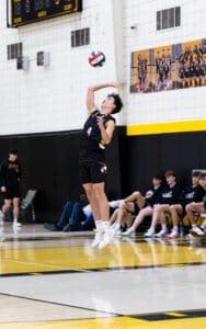 A volleyball player jumps to serve during a match, with teammates and a scoreboard visible in the background.