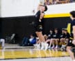 Volleyball player jumps to serve in a competitive match with teammates on the sidelines and a scoreboard visible.