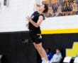 High school volleyball player jumping to serve the ball during a game, with teammates visible in the background.