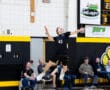 High school volleyball player jumps to serve a volleyball during a game, with spectators in the background.