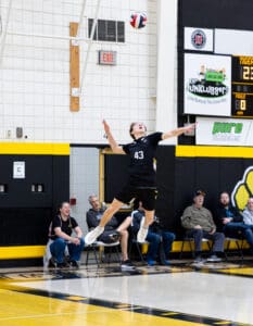 High school volleyball player jumps to serve a volleyball during a game, with spectators in the background.