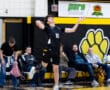 A high school volleyball player serves a ball during a game, with spectators watching in the background.