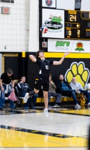 A high school volleyball player serves a ball during a game, with spectators watching in the background.