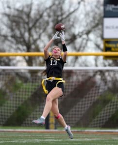 Girl in flag football gear jumps to catch a football, showcasing athleticism on a field with goalposts in the background.