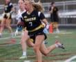 Female athlete wearing a black and yellow jersey runs with a football on a field during a flag football game.
