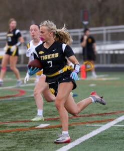 Female athlete wearing a black and yellow jersey runs with a football on a field during a flag football game.