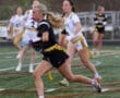 A female flag football player runs with the ball as teammates pursue her on a green field during a game.