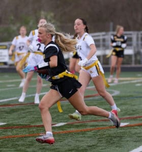 A female flag football player runs with the ball as teammates pursue her on a green field during a game.