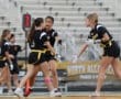 Three female athletes in black jerseys and yellow belts celebrate on the field during a flag football game.
