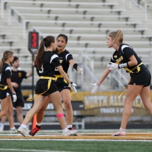 Three female athletes in black jerseys and yellow belts celebrate on the field during a flag football game.