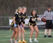 Three female athletes in black jerseys prepare for a play in a flag football game, with a referee observing nearby.