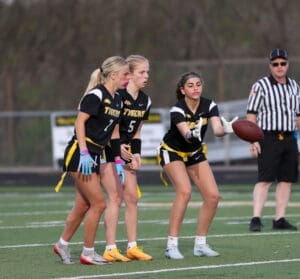 Three female athletes in black jerseys prepare for a play in a flag football game, with a referee observing nearby.