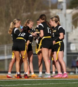 A group of female athletes in black uniforms discussing strategy during a flag football game on a sunny day.