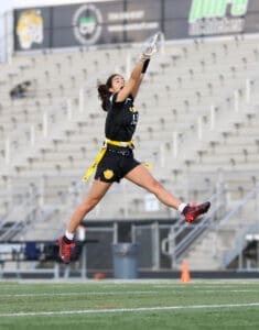 Female athlete leaping to catch a flag football pass, wearing a black jersey and yellow flags on a football field.