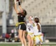 Girl in black uniform jumps to catch a football in a flag football game, while opposing players try to block her.