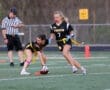 Two female flag football players in black jerseys strategizing on the field, with a referee observing the game.