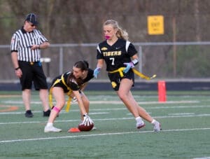 Two female flag football players in black jerseys strategizing on the field, with a referee observing the game.