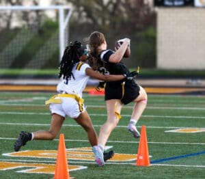 Two young girls playing flag football, one running with the ball while the other attempts to pull her flag.