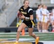 Female athlete in a black football uniform running with a football on the field during a game. Energized action shot.
