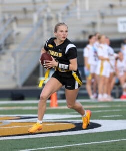 Female athlete in a black football uniform running with a football on the field during a game. Energized action shot.