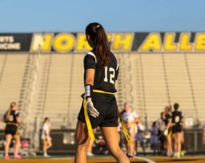 Female flag football player in black jersey, number 12, prepares for the game with North Allegheny stadium in the background.