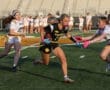 Girl in black jersey runs with football, evading defenders in a vibrant flag football game on a sunny field.