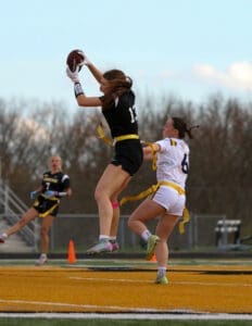 Girl in black jersey jumps to catch a football while another player in white attempts to block her during a game.