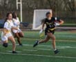 A girl in a black football jersey runs with the ball, evading two opponents on the field during a game.