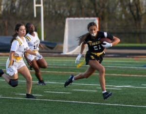 A girl in a black football jersey runs with the ball, evading two opponents on the field during a game.