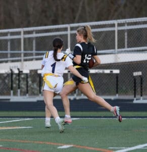 Two female athletes compete in a flag football game, one attempting to evade the other while holding the ball.