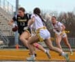 A female football player in a black jersey runs with the ball, pursued by teammates on the field during a game.