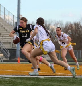 A female football player in a black jersey runs with the ball, pursued by teammates on the field during a game.
