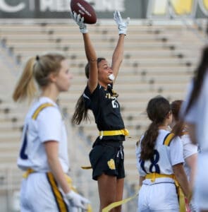 Girl celebrating a successful play in flag football, raising her arms in excitement while teammates observe.