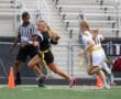 Female athlete running with a football in flag football game, referee observing, colorful uniforms on a turf field.