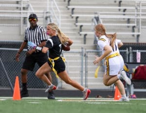Female athlete running with a football in flag football game, referee observing, colorful uniforms on a turf field.