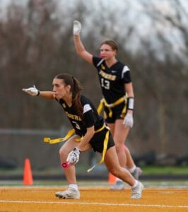 Two female flag football players in black uniforms signal during a game, demonstrating teamwork and athleticism on the field.