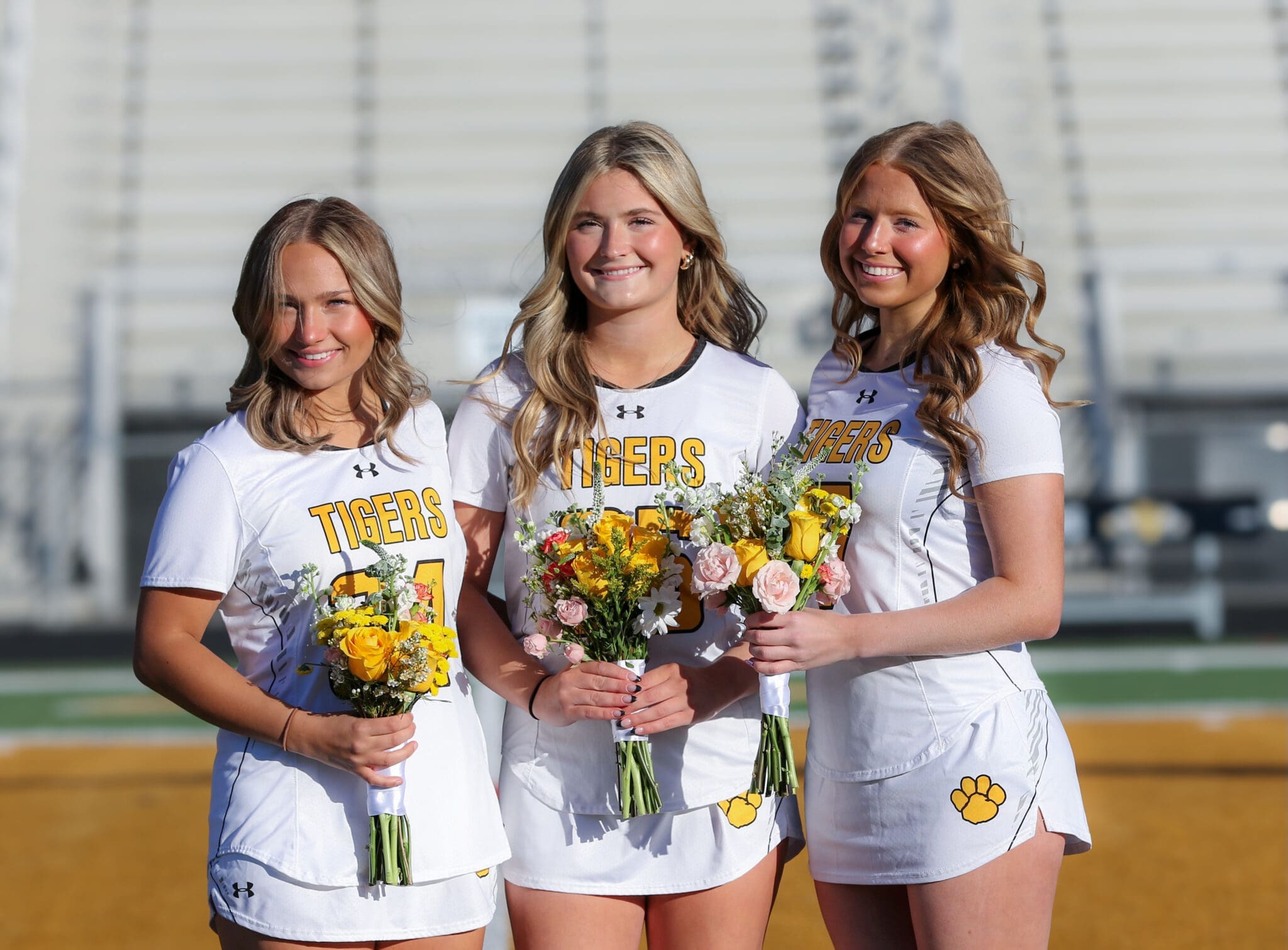 Three female lacrosse players from the Tigers team, wearing jerseys and holding floral bouquets, smiling together.