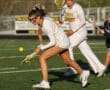 High school girls' lacrosse players competing for the ball on a sunny day during a match on a green turf field.