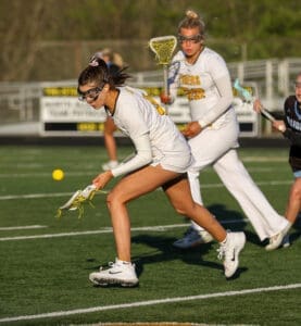 High school girls' lacrosse players competing for the ball on a sunny day during a match on a green turf field.
