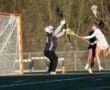 Girls' lacrosse match action: a player shoots while the goalie attempts to block the shot in bright daylight.