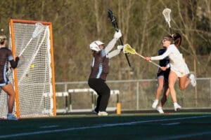Girls' lacrosse match action: a player shoots while the goalie attempts to block the shot in bright daylight.