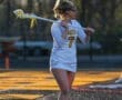 Female lacrosse player in a white uniform with the number 7, holding a stick on a field during practice at sunset.