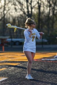 Female lacrosse player in a white uniform with the number 7, holding a stick on a field during practice at sunset.
