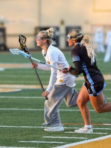 Two female lacrosse players sprint on the field, focused on the game, showcasing teamwork and athleticism.
