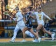 Three lacrosse players compete on the field during a game, showcasing action and athleticism against a sunset backdrop.
