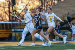 Three lacrosse players compete on the field during a game, showcasing action and athleticism against a sunset backdrop.