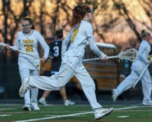 Female lacrosse players in white uniforms sprinting on the field at sunset, showcasing athleticism and teamwork.