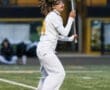 Female lacrosse player in white jersey and sweatpants, preparing for a play on the field with a yellow lacrosse stick.